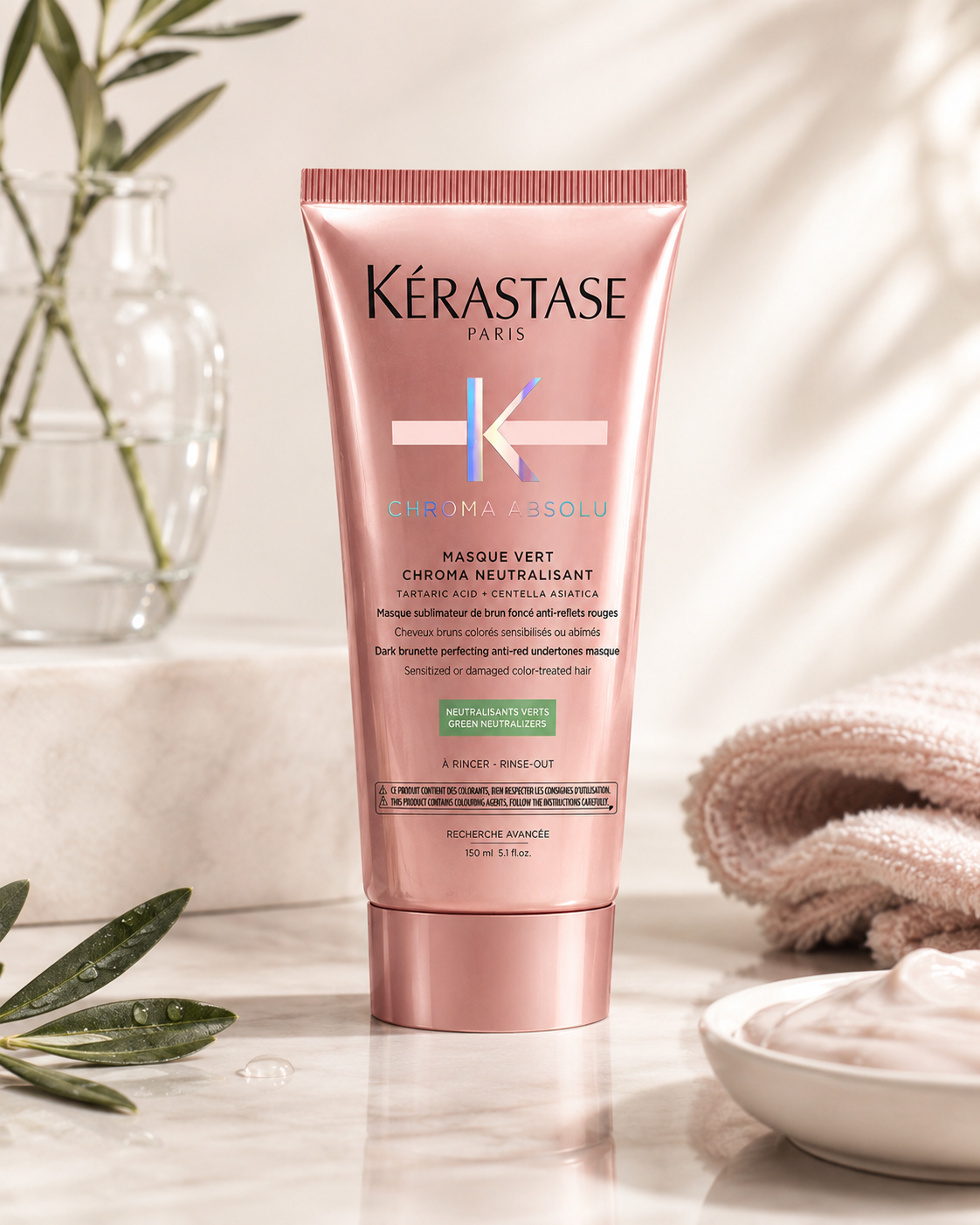 Kérastase Chroma Absolu Masque Vert Chroma Neutralisant elegantly displayed on a polished white marble vanity in warm natural daylight. The premium haircare scene features fresh olive branches in a clear glass vase, neatly folded blush-colored spa towels, a ceramic bowl containing creamy hair mask, subtle water droplets on the marble surface, and soft botanical shadows creating an elegant salon-spa atmosphere.